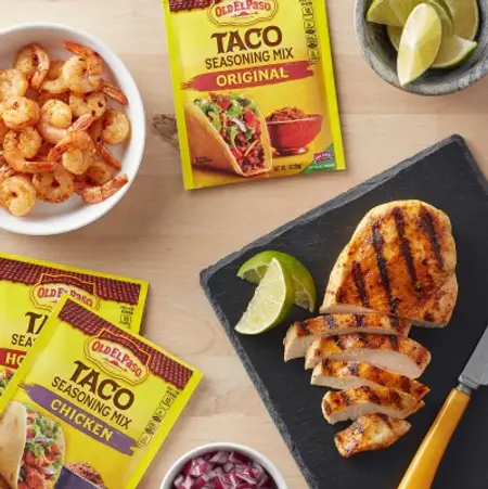 Three packets of Old El Paso Taco Seasoning on a wooden table with shrimp and grilled chicken